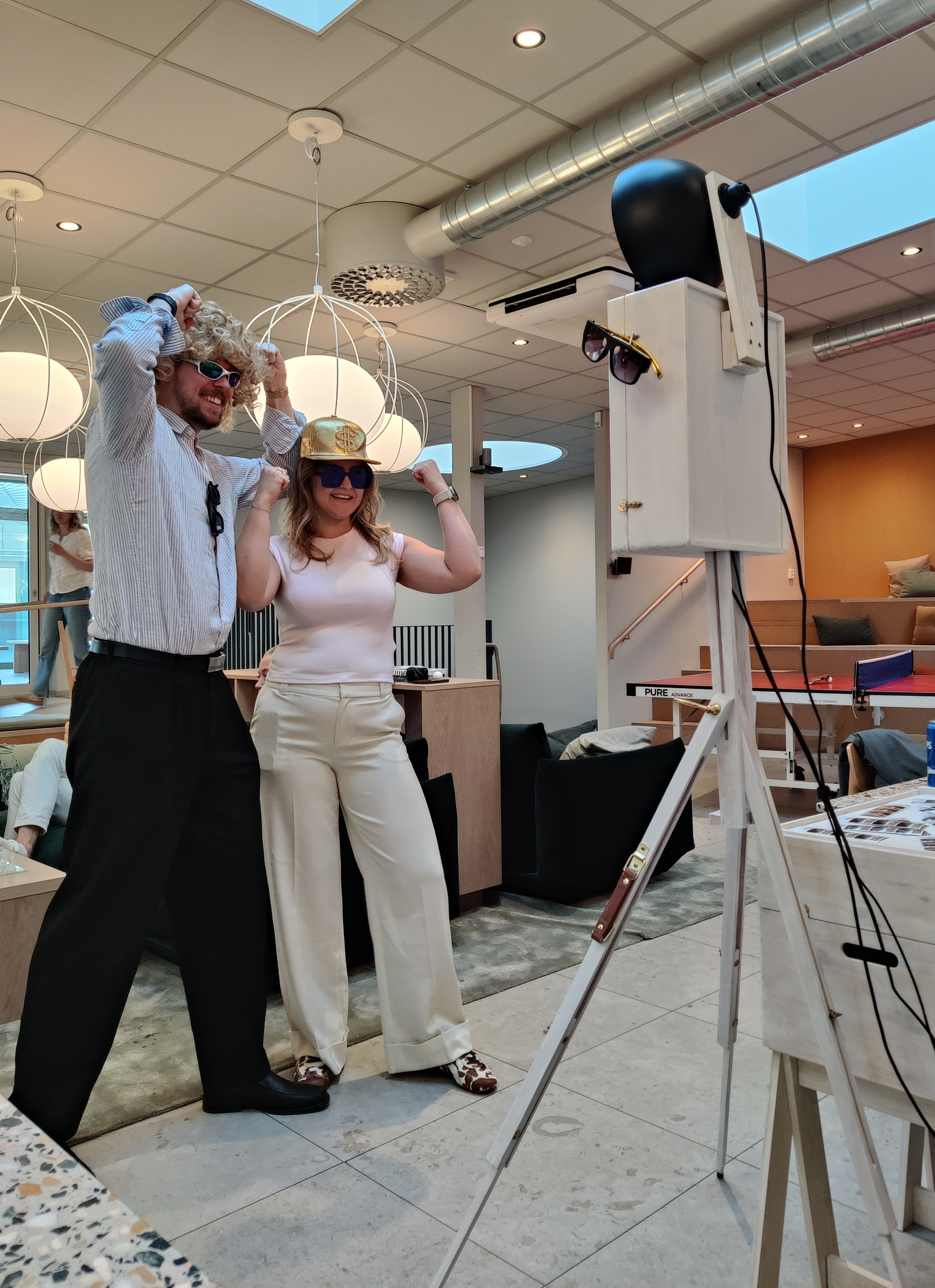 Två kollegor i peruk och guldkeps poserar framför Mabooths fotobås på ett företagsevent