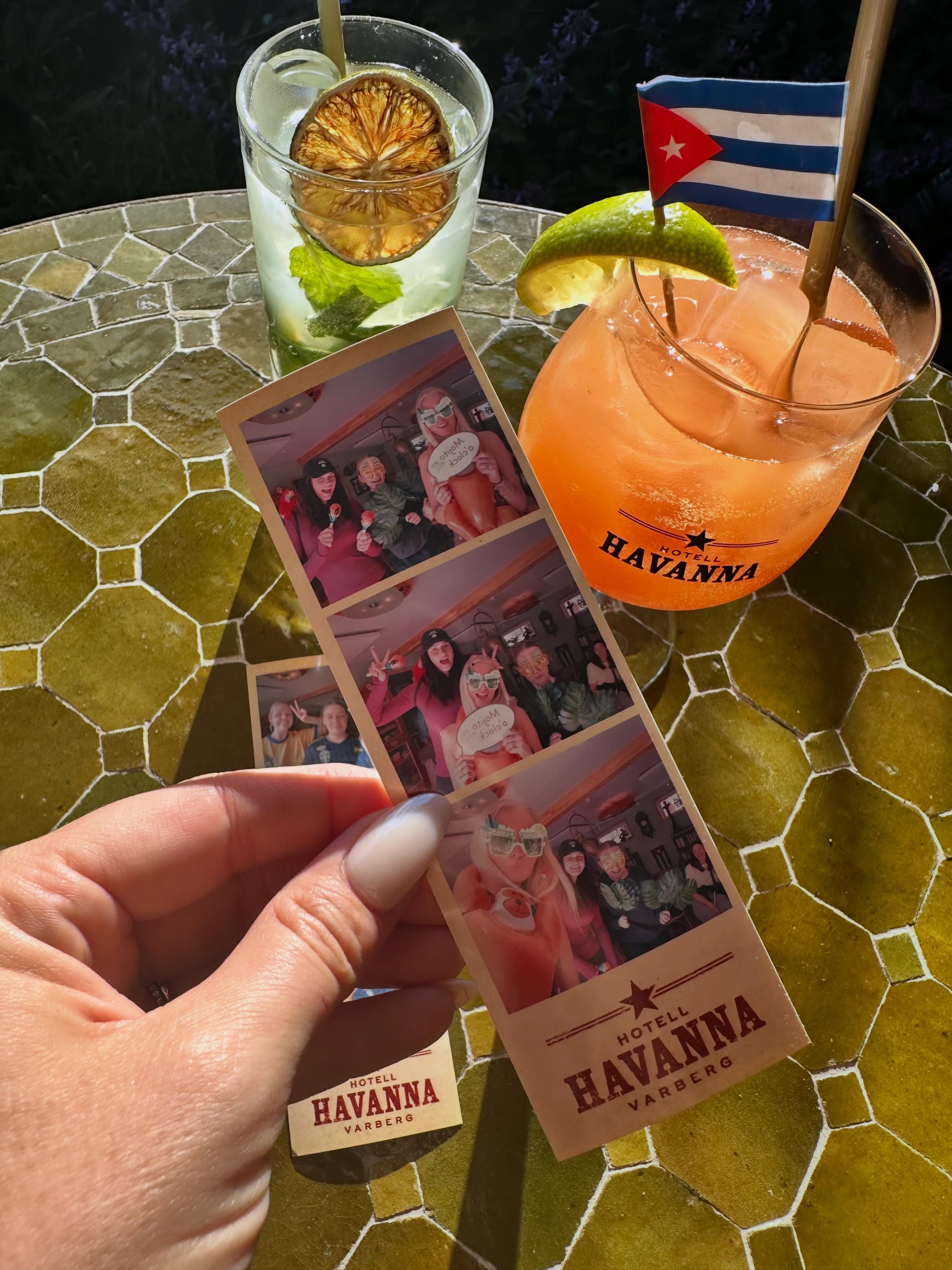 Hand som håller upp en nyutskriven fotoremsa från Mabooth bredvid cocktails på Hotell Havanna i Varberg
